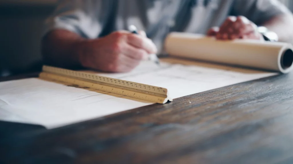 Un professionnel réalise un dessin technique sur des plans à l’aide d’une règle.